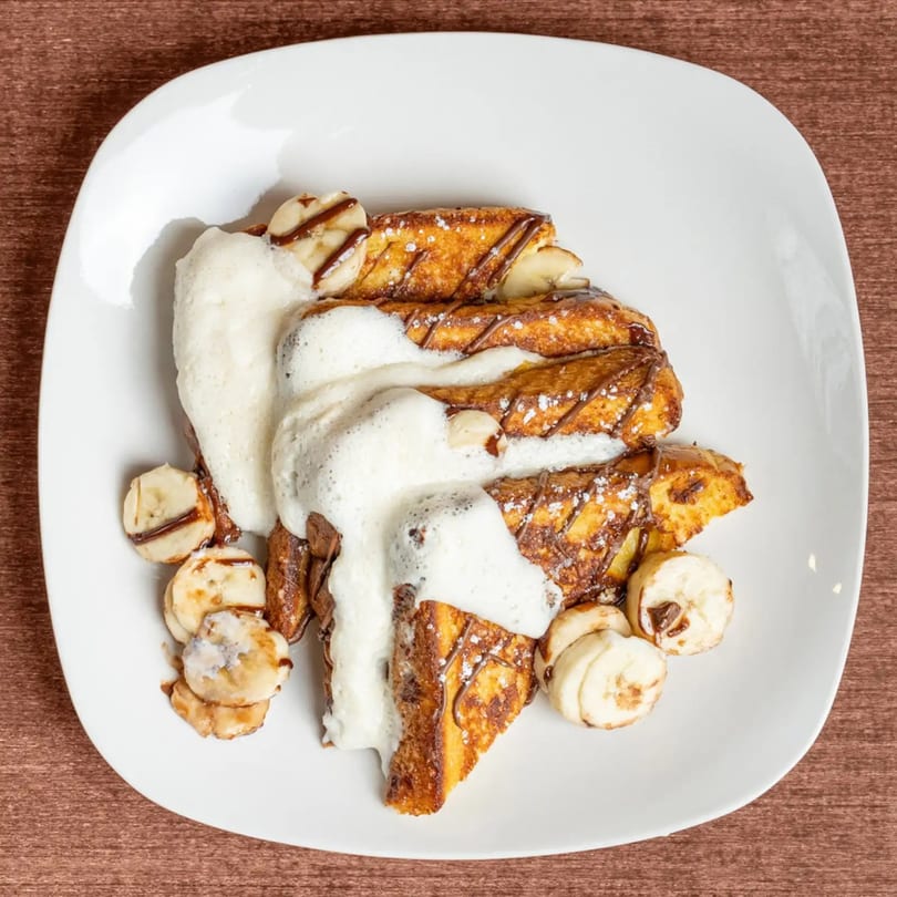 Golden French toast topped with whipped cream, chocolate drizzle, powdered sugar, and banana slices.