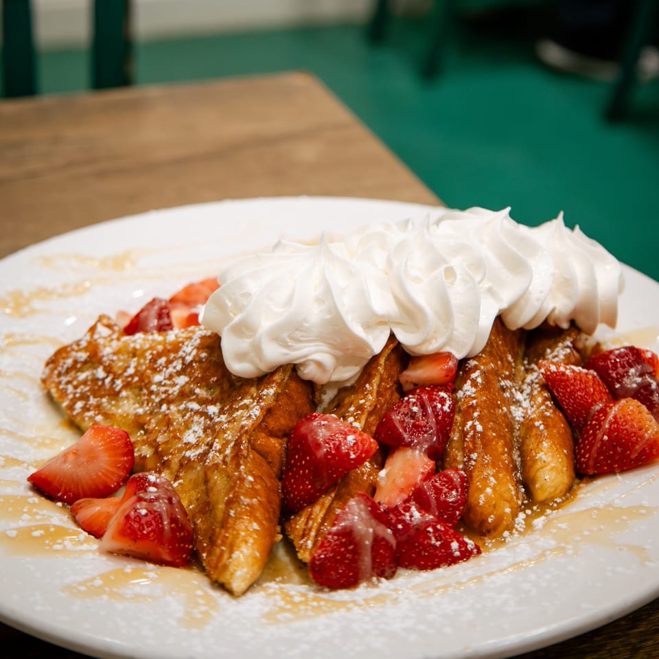 Golden French toast topped with whipped cream, fresh strawberries, powdered sugar, and syrup.
