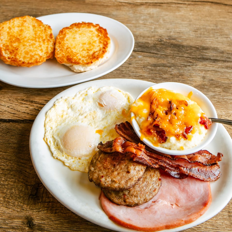 A hearty breakfast plate with fried eggs, sausage, ham, bacon, cheesy grits, and two biscuits.