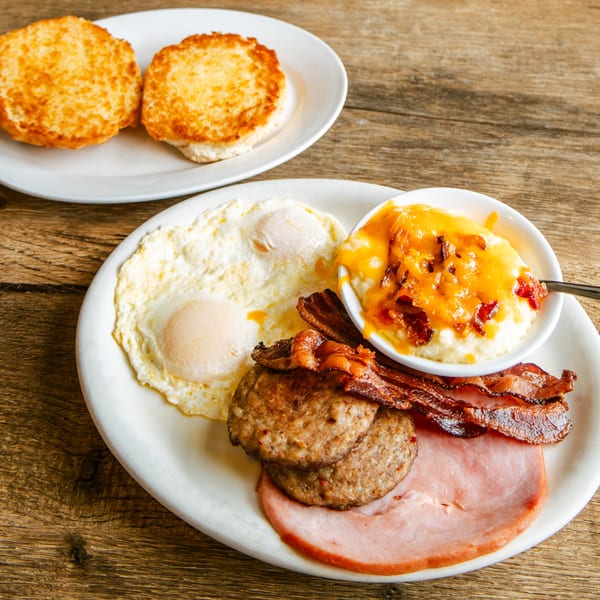 A full breakfast plate with eggs, bacon, sausage, ham, cheesy grits, and two biscuits.
