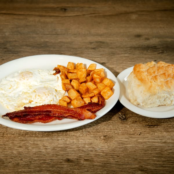 Breakfast plate with fried eggs, crispy bacon, home fries, and a biscuit on a wooden table.
