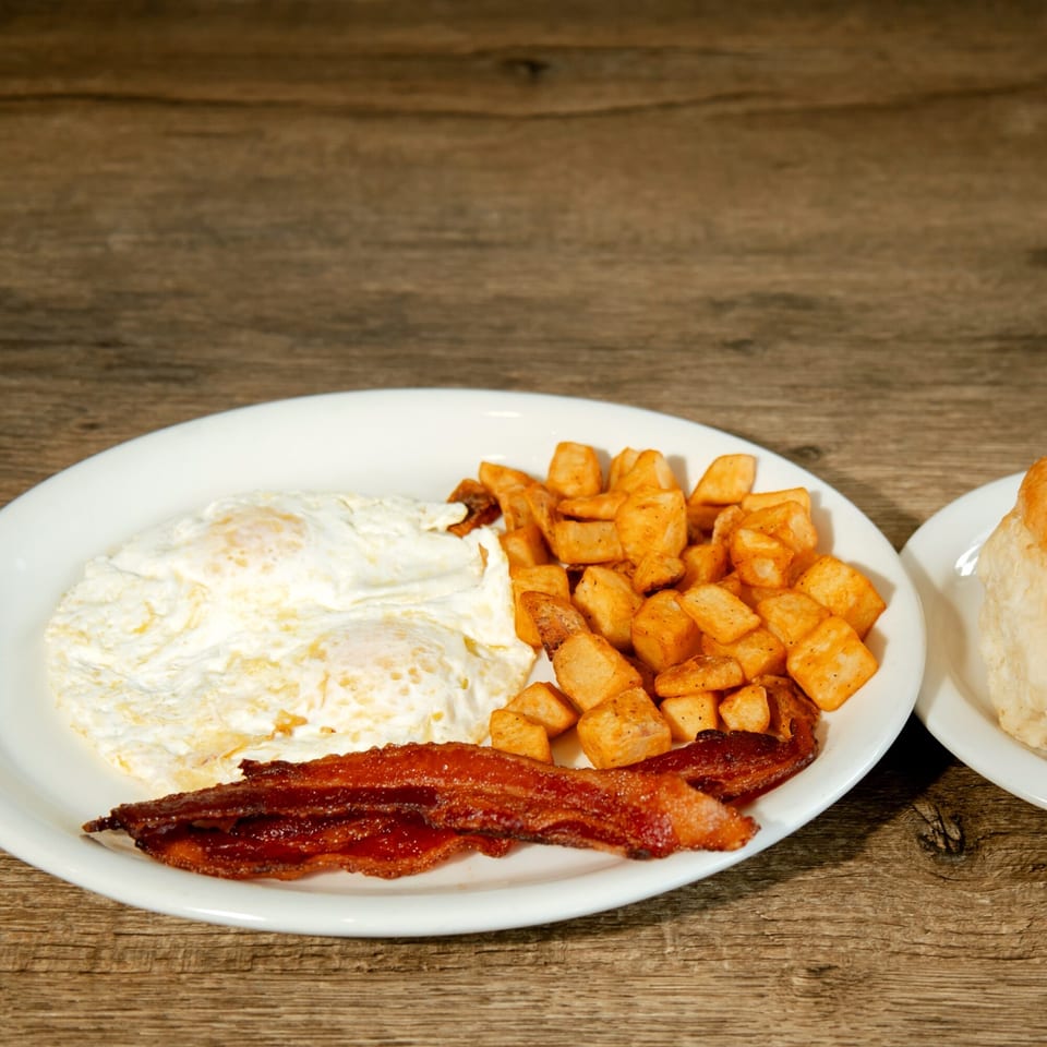 A breakfast plate with fried eggs, crispy bacon, and seasoned home fries on a wooden table.