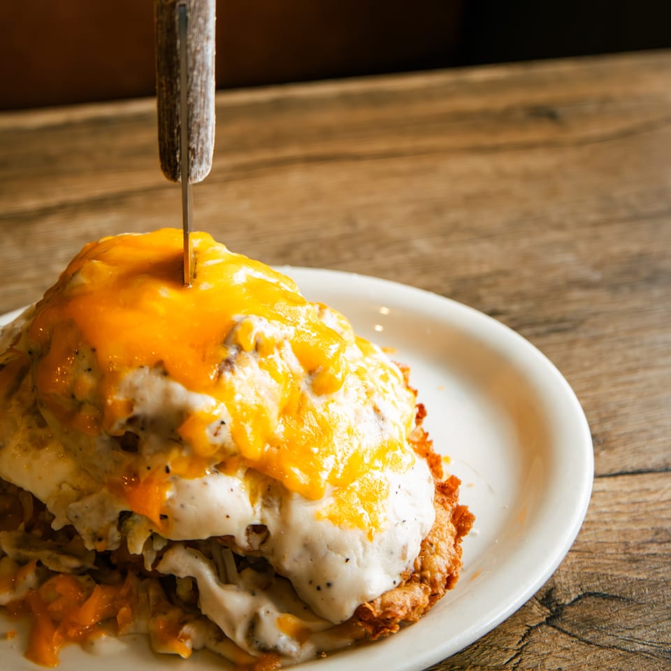 A knife piercing a stack of hash browns, gravy, and melted cheese on a white plate.