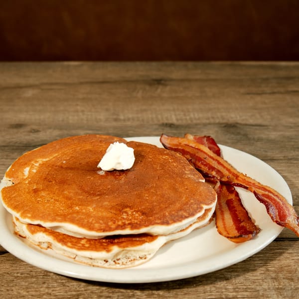Stack of pancakes with butter and crispy bacon on a white plate on a wooden table.