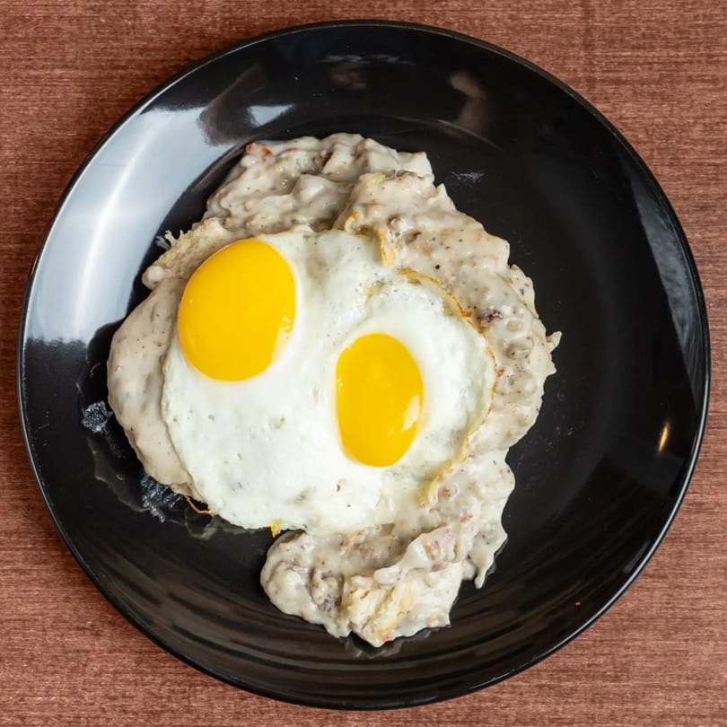 Two sunny-side-up eggs on a bed of creamy white gravy and biscuits, served on a black plate.