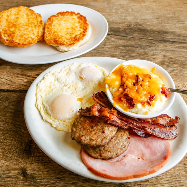 A hearty breakfast platter with eggs, bacon, sausage, ham, cheesy grits, and two biscuits.