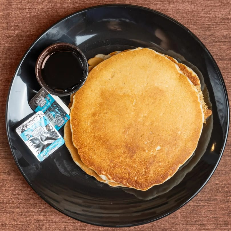 A stack of golden pancakes with syrup and two butter packets on a black plate.