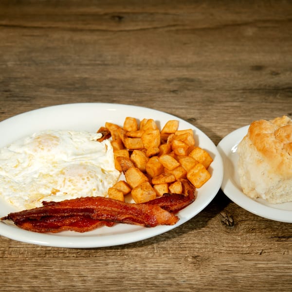 A breakfast plate with fried eggs, crispy bacon, and home fries, with a biscuit on a side plate.
