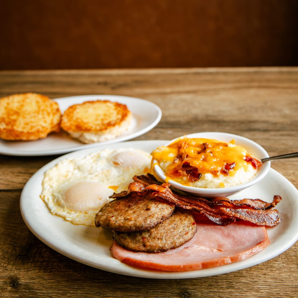 Hearty breakfast with fried eggs, bacon, sausage, ham, cheesy grits, and two biscuits on a wooden table.