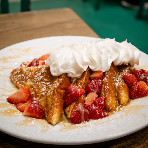 Golden French toast topped with whipped cream, fresh strawberries, powdered sugar, and syrup.