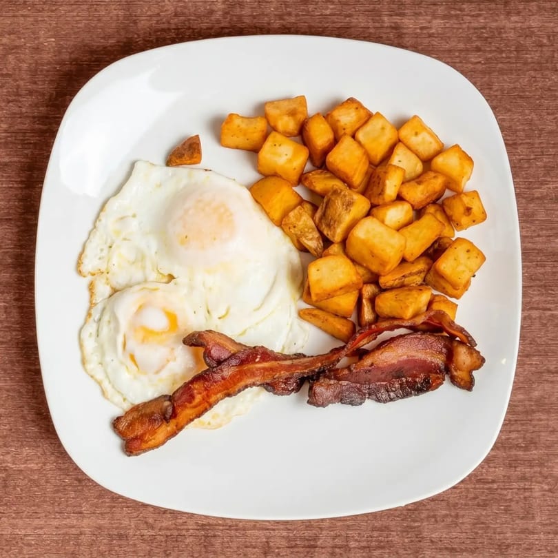 A breakfast plate with two fried eggs, crispy bacon strips, and golden-brown home fries.