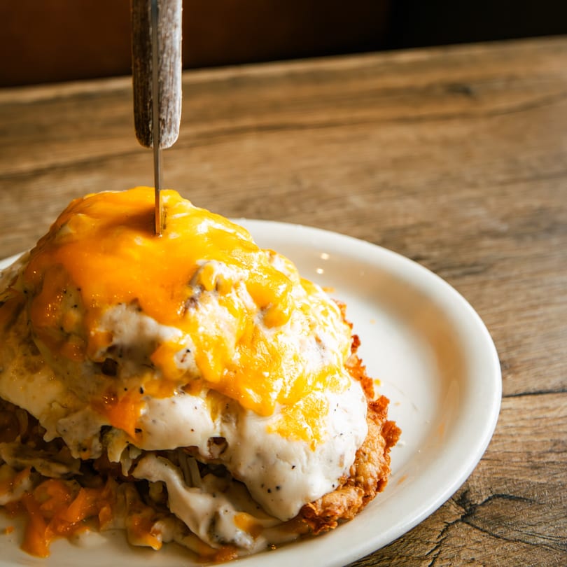 A knife stuck into a stack of hash browns, gravy, and melted cheese on a white plate.