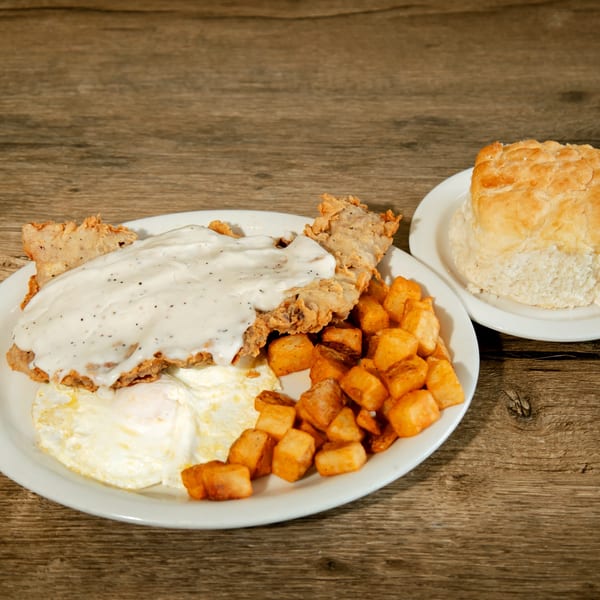 Chicken fried steak with gravy, fried eggs, home fries, and a biscuit on a wooden table.