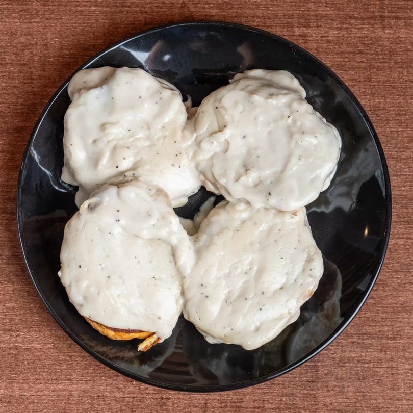 Four biscuits covered in creamy white gravy with black pepper on a black plate.