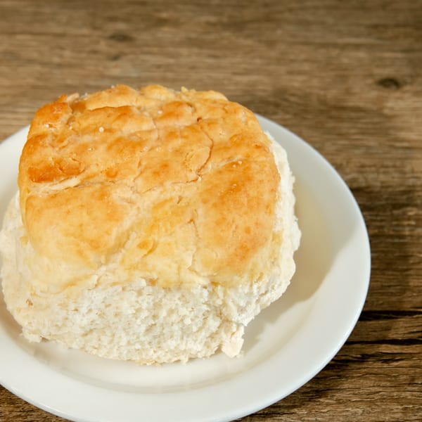 A golden-brown biscuit on a white plate, resting on a rustic wooden table.
