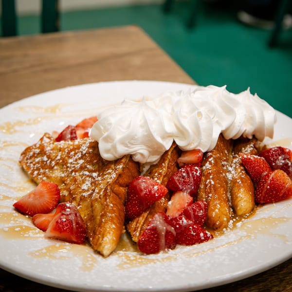 Golden French toast topped with whipped cream, fresh strawberries, powdered sugar, and syrup on a white plate.
