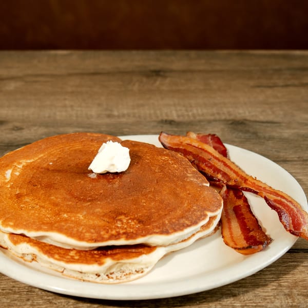Stack of golden pancakes with a dollop of butter and crispy bacon strips on a white plate.