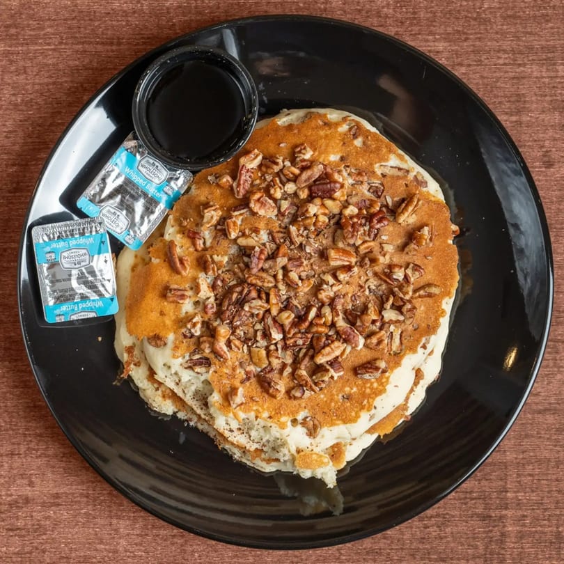 Pancakes topped with pecans, served with syrup and two packets of whipped butter on a black plate.